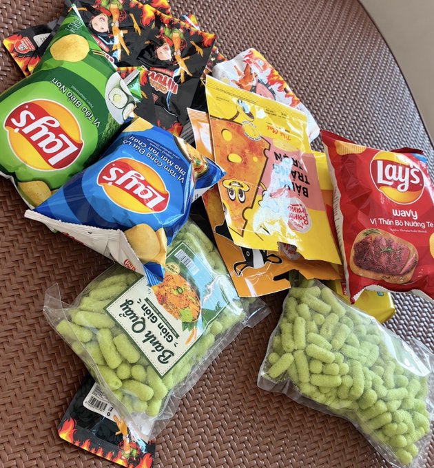 A collection of assorted snack packages including Lay's chips in green, blue, and red bags, yellow Banana-themed packaging, and two clear bags of green pea snacks labeled 'Banh Que' scattered on a brown woven mat.