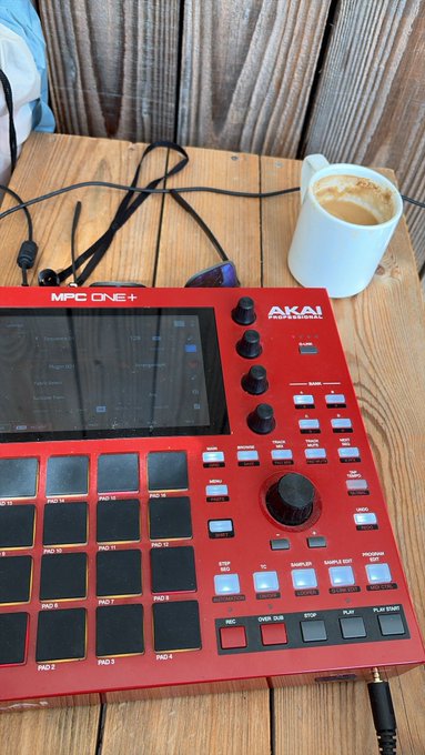 A red AKAI MPC One+ drum machine sits on a wooden surface next to a white mug of coffee.