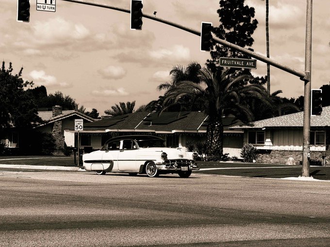 A vintage lowrider cruises down a quiet street beneath a "FRUITVALE AVE" sign, with a 50 mph speed limit sign nearby, capturing a nostalgic moment that fits the artistic, photography-based NFT "Passing Through" by @Sandytoes2211 on objkt.com.