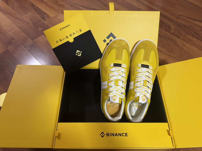 A pair of limited-edition Binance-branded yellow sneakers, accompanied by a protective bumper case and a card with "Binance" branding and Chinese characters, unboxed in bright yellow packaging on a wooden floor as a Mid-Autumn Festival gift.