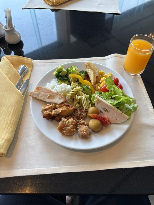 A white plate filled with a diverse buffet selection including fried chicken, rice, broccoli, salad, pasta, sausage, olives, and tomatoes sits on a white placemat beside a glass of orange juice on a dark table.