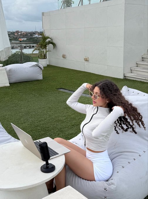 A woman with curly hair lounges on a white bean bag on a rooftop terrace, wearing a white long-sleeve top and skirt while using a laptop, with a microphone and small table nearby overlooking a coastal view. The "Gm & happy Saturday" caption fits the laid-back weekend vibe of someone enjoying a relaxed morning outdoors while working or browsing on their laptop in a scenic setting.