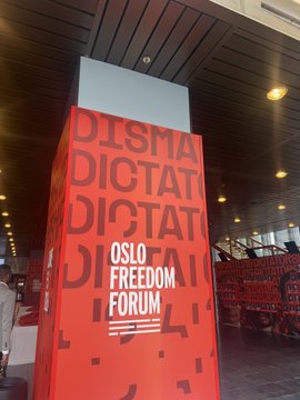 The image features a tall, red pillar with white text that reads "OSLO FREEDOM FRONT" at the top of the pillar. Below the pillar, there are two people sitting at tables, possibly attending an event or gathering related to the freedom front. The room is filled with various objects such as chairs and dining tables scattered throughout the space.