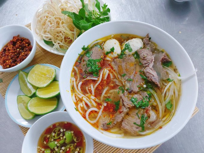 A bowl of Vietnamese pho noodle soup with beef and herbs, accompanied by side dishes of bean sprouts, lime wedges, and chili condiments.