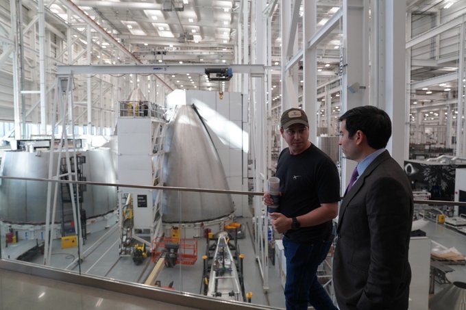 Two men, one in a black t-shirt and cap and the other in a suit, stand on a balcony overlooking a large industrial facility with a white spacecraft component being assembled. This image relates to the post as it showcases the impressive, movie-like setting of Starbase, SpaceX's facility for building the Starship rocket.