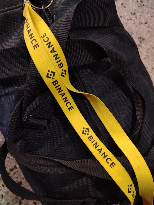 A black duffel bag with a yellow lanyard bearing the repeating text 'BINANCE' and the Binance logo.