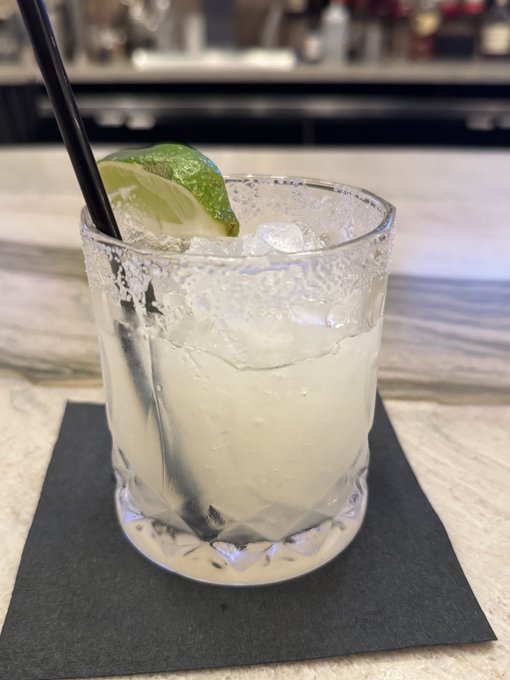 A photo of a clear cocktail (likely a margarita or gin and tonic) with ice and a lime wedge, served in a salt-rimmed rocks glass with a black straw on a black napkin.