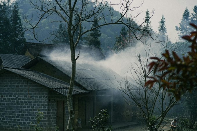 The image shows an old brick building with smoke coming out of its chimney, surrounded by trees. The scene appears to be set in a forested area or near a mountain range. There are two people present in the image - one person is standing closer to the left side and another person is located more towards the right side of the frame.