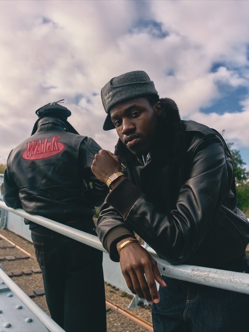 A young man wearing a grey cap and black leather jacket poses while leaning against a railing, with another person behind him wearing a jacket reading 'Waileks'.
