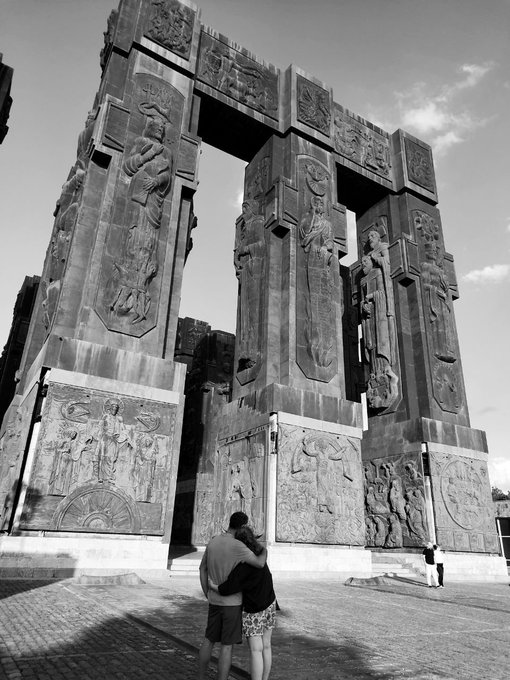 A black and white photograph of a couple embracing in front of a massive, intricately carved stone monument featuring religious figures in arched alcoves.