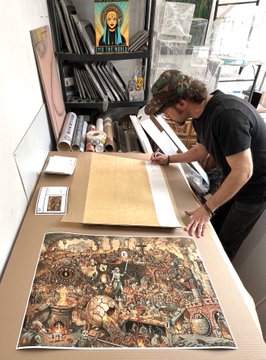 An artist with curly hair works at a desk with detailed artwork, surrounded by rolled prints and storage shelves containing a small religious icon.