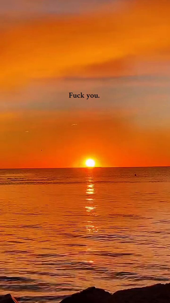 The image features a sunset over the ocean with the text "FUCK YOU" written above it. The sun is setting behind the horizon, casting an orange glow across the sky. In the foreground of the photo, there are rocks visible on the beach.