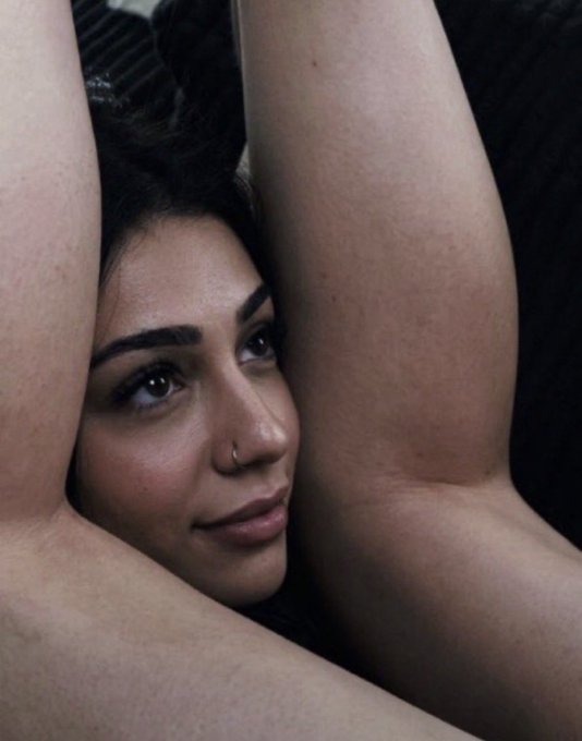 An artistic close-up photograph of a woman with dark hair and a nose ring, framed by her raised arms, looking upward with a contemplative expression.