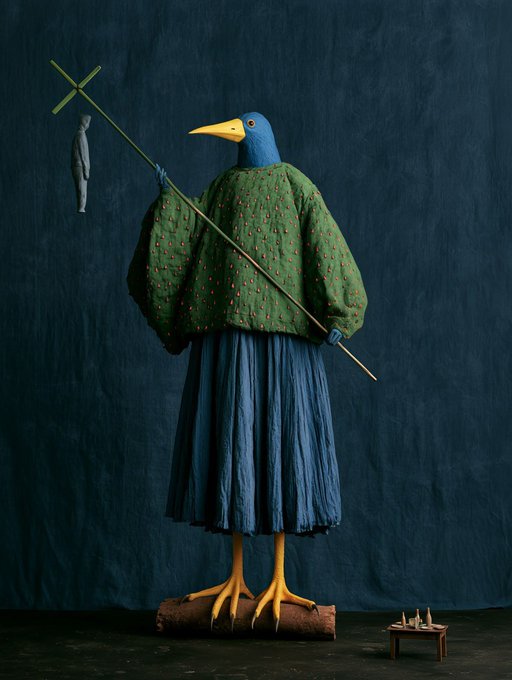A surreal sculptural figure with a blue bird's head, yellow beak, and bird legs stands atop a log, wearing a green patterned sweater and long blue pleated skirt while holding a long staff with a small clay figure hanging from it, set against a dark navy backdrop with a tiny desk and figures to the side—evoking the mysterious, bureaucratic guardian described in the caption whose purpose remains unknowable and whose offerings go untouched.