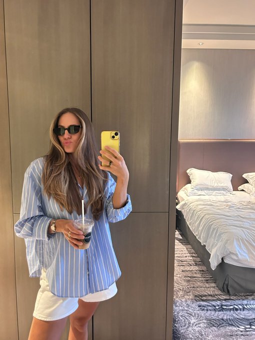 A woman takes a mirror selfie in a bedroom holding a phone and an iced coffee, wearing a blue and white striped button-down shirt, white shorts, and sunglasses.