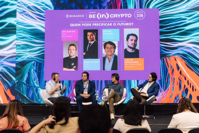 Four men sit on a stage during a panel discussion beneath a screen labeled 'BINANCE BE [IN] CRYPTO' and 'PAÇO 23B' featuring speakers Roy Shahaf, Arthur Laçache, João Accoli, and Luis de Macedo Soares.