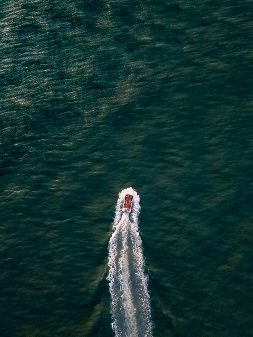 An aerial black-and-white photograph of a small red boat moving through deep blue-green water, leaving a long white wake behind it.