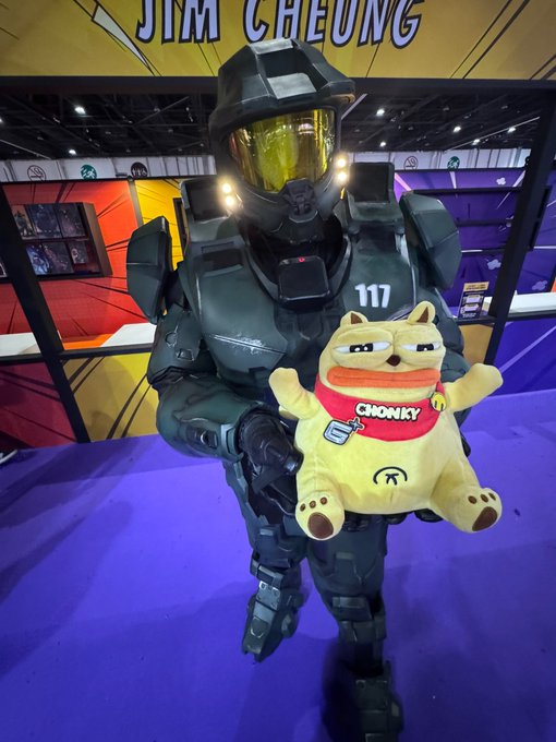 A person in a Master Chief Halo costume (number 117) holding a 'CHONKY' Shiba Inu plush toy at what appears to be a convention.