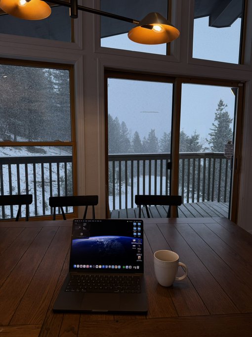 A laptop displaying a stock or crypto trading chart sits on a wooden table next to a coffee mug, with a snowy, overcast forest view through the large windows behind it. The bleak, isolated scene perfectly captures the post's sentiment of feeling stranded and empty-handed after putting faith in cryptocurrency investments.