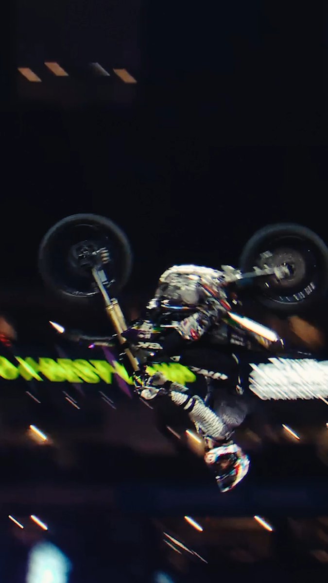 An action shot of a motocross rider performing an aerial backflip trick on a dirt bike during a nighttime event, with motion-blurred stadium lights and yellow text in the background.