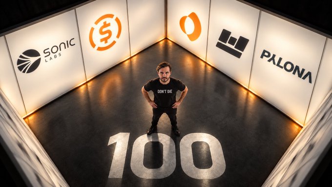 A man wearing a 'DON'T DIE' t-shirt stands in a box-like room with logos for Sonic Labs, OKX, and Palona displayed on the walls, with the number '100' painted on the floor beneath him.