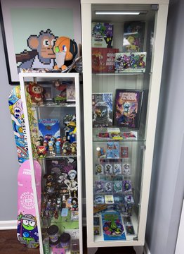 A tall glass display cabinet filled with video game collectibles and figures, with a skateboard featuring colorful graphics leaning against its side.