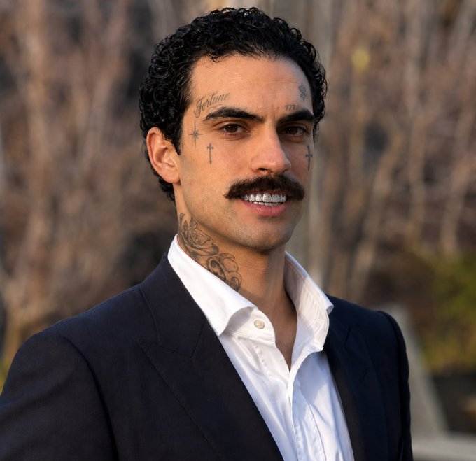 A portrait of a man with curly dark hair, a mustache, diamond-encrusted teeth, and face/neck tattoos, wearing a black blazer over a white shirt.
