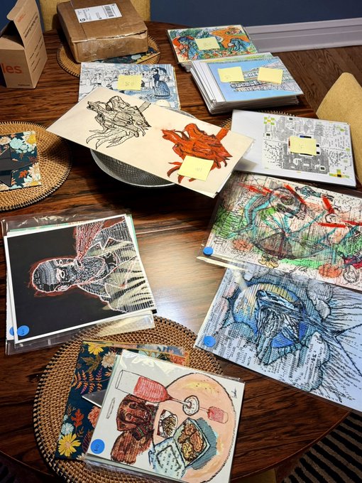 A wooden table is covered with various pieces of artwork and illustrations, including drawings of a dragon, people, and food, some with yellow sticky notes attached.