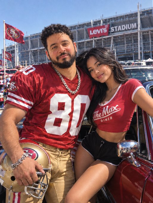 A man wearing a San Francisco 49ers jersey with the number 81 holds a football helmet, standing with a woman in a red 'The Max Diner' crop top in front of Levi's Stadium.