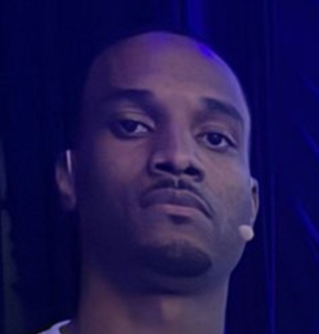 A close-up portrait of a man with a serious expression wearing a small earpiece microphone against a dark blue background, likely from a televised interview or event.