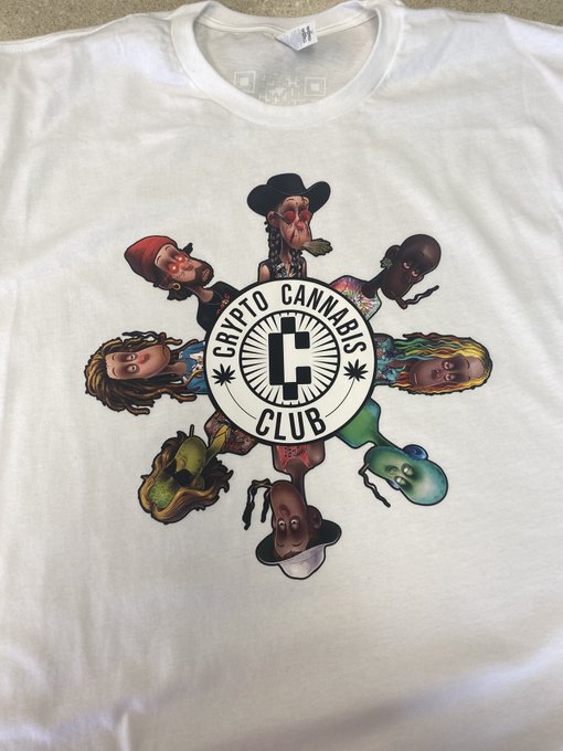 A white t-shirt featuring a 'Crypto Cannabis Club' logo with a stylized 'C' in the center, surrounded by cartoon character heads with cannabis-themed imagery arranged in a sunburst pattern.