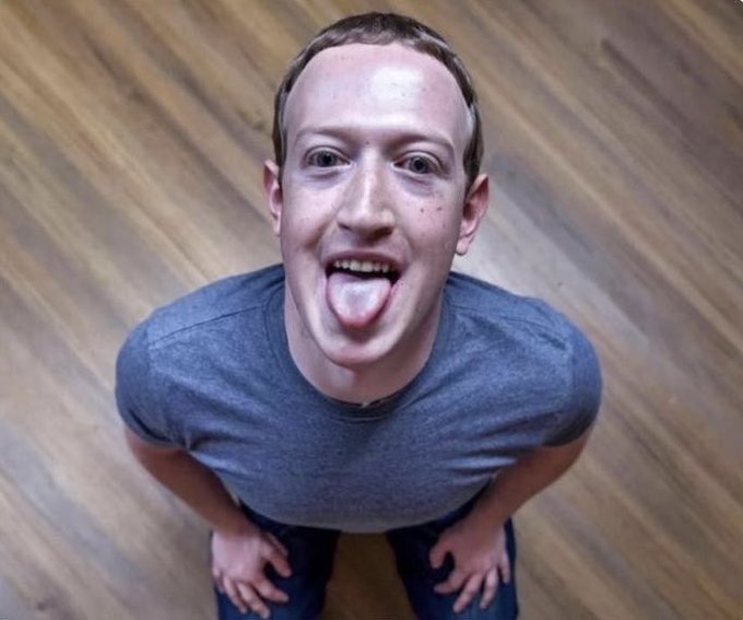 A fisheye-lens photo of Mark Zuckerberg in a gray t-shirt, kneeling on a wooden floor and sticking his tongue out playfully at the camera.