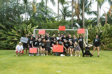 A large group of people wearing matching black outfits pose on a lush green lawn in front of a soccer goal. Several people in the group are holding up red signs with white text, with 'RFG' partially visible.