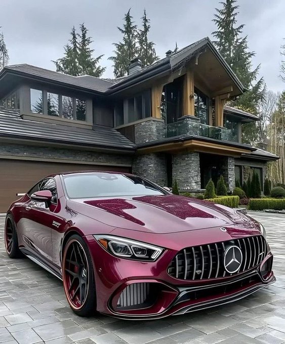 A burgundy red Mercedes-AMG coupe parked on a paved driveway in front of a large modern stone and wood house surrounded by trees.