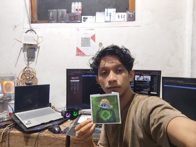 A young man takes a selfie at his workstation with multiple monitors and a laptop, holding up a printed photo of a cartoon character wearing a cap with a design resembling an Alpaca logo.