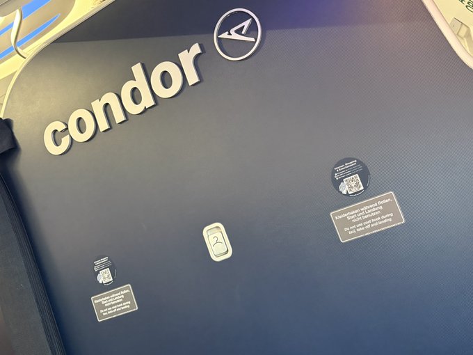 A Condor airline seatback displays the brand's logo and signage in German regarding the "Condor-Spass" children's program and power outlets, taken from a passenger's seat during their return flight home.