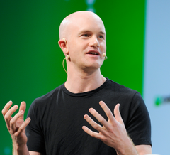 A bald man wearing a black t-shirt speaking with his hands gesturing, appearing to be giving a presentation on a stage with a green background.