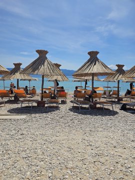 The image shows a beach scene with several umbrellas providing shade for people sitting or relaxing on chairs, as well as others walking along the shoreline. The beach appears to be empty at the moment, giving an impression of tranquility and relaxation. The photo is taken from a low angle, which emphasizes the height of the umbrellas and adds depth to the scene. The overall atmosphere seems calm and inviting, with people enjoying their time by the water under the shade provided by the numerous umbrellas.