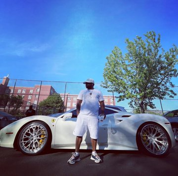 A man wearing all white, including a t-shirt, shorts, sneakers, and a cap, stands next to a white sports car on a sunny day with a chain-link fence and trees in the background.