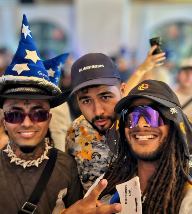 A selfie of three smiling men at what appears to be a sporting event; the man on the left wears a blue wizard hat and sunglasses, the middle man wears a "RAFERSKICKINPS" cap, and the man on the right sports a Cleveland cap, purple sunglasses, and dreadlocks.