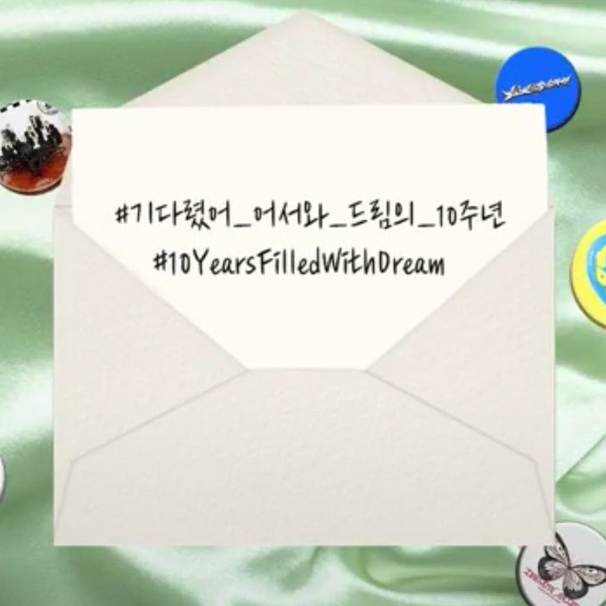 A handwritten note inside an envelope reading '#기다렸어_아시라_드림의_10주년 #10YearsFilledWithDream', placed on a light green fabric with decorative buttons.