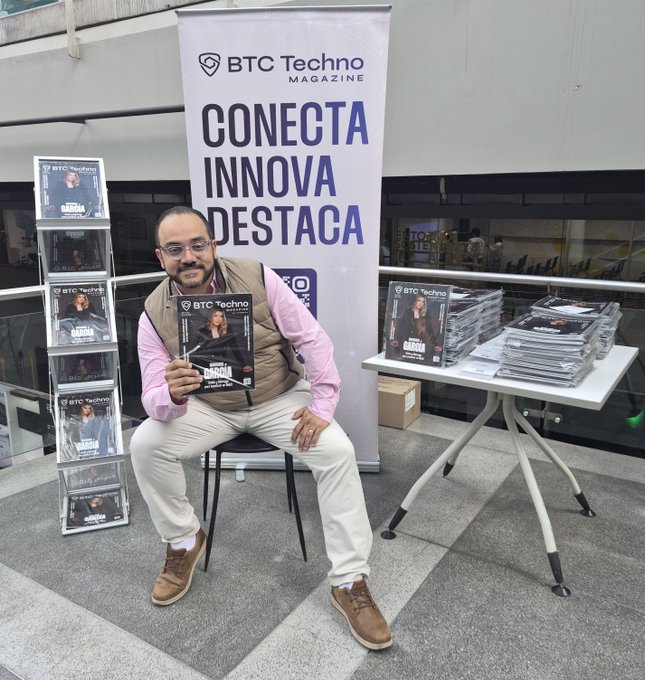 A man in a pink shirt and tan vest sits holding a BTC Techno Magazine, posing in front of a banner reading 'CONECTA INNOVA DESTACA' with magazine racks on either side.