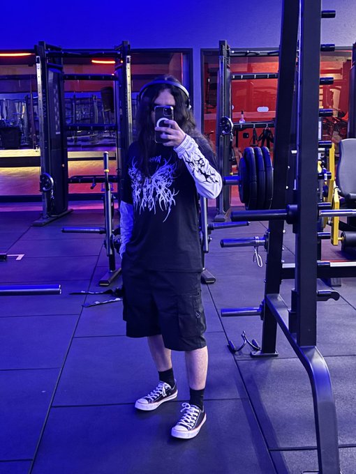 A mirror selfie of a person in a black graphic long-sleeve shirt and shorts, wearing headphones and taking the photo with a phone in a gym with purple lighting.