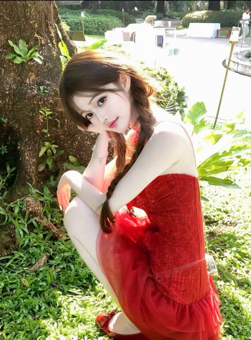 A young woman in a red dress crouching by a tree in a garden setting.