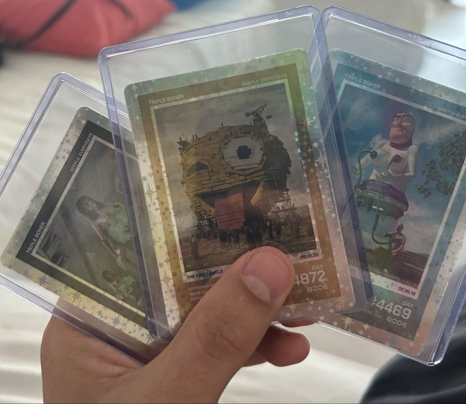 A hand holds three Beeple "Triple Bonner" collectible cards in protective sleeves, each marked "WORLD EVENT SERIES" with dates (09.03.20, 07.06.20) and edition numbers (#4872 and #4469), featuring Beeple's signature surreal digital art style. The cards illustrate the post's mention of collecting these limited-edition pieces, which have become the user's new collecting obsession.
