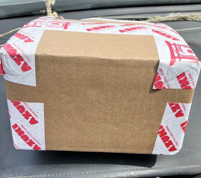 The image shows a brown cardboard box with red and white stripes, sitting on top of a black surface. The box appears to be a delivery or shipping container, possibly containing a new item or product. The text "First package arrived! This is just my booster box of @AzukiTCG still waiting my case and starters!" suggests that the box has been recently received and is now being prepared for use.
