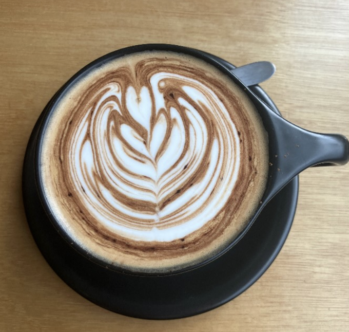 A black cup of coffee viewed from above features intricate latte art in the shape of a leaf or rosetta pattern, resting on a wooden table.