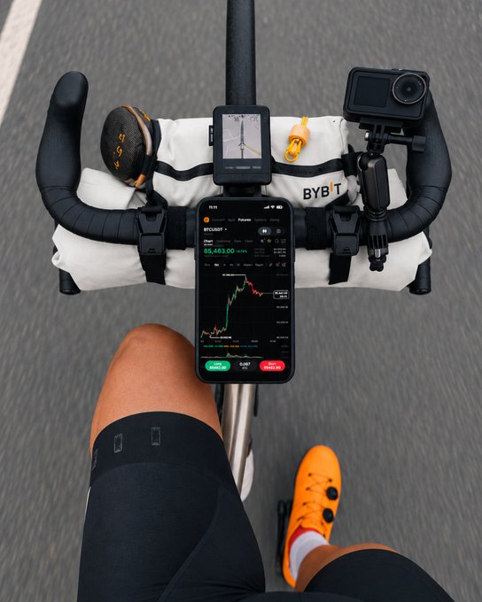 A first-person view from a cyclist shows a handlebar bag with "BYBI" branding, holding a GPS cycling computer and a GoPro, while a phone mounted below displays a BTCUSD chart trading at 116,863.00, capturing the post's moment of pausing to check crypto markets mid-ride.
