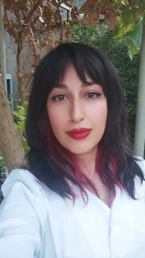 A selfie of a woman with dark hair featuring pink highlights, wearing a nose ring, red lipstick, and a white blouse, posed against greenery.