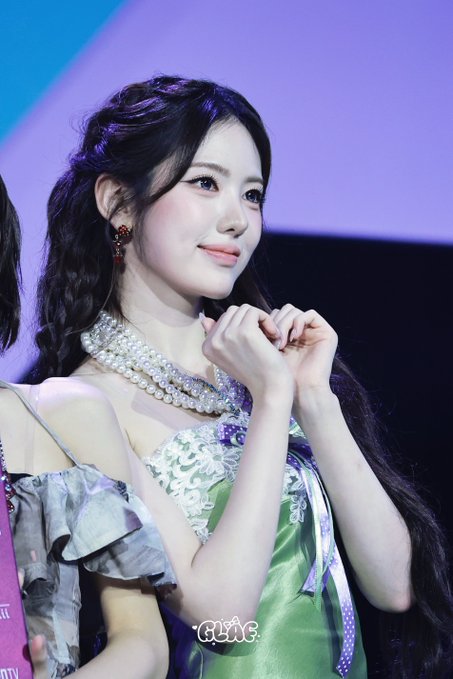 A photograph of a young woman on stage, wearing a green dress with pearl detailing and a small red earring, smiling with her hands clasped.